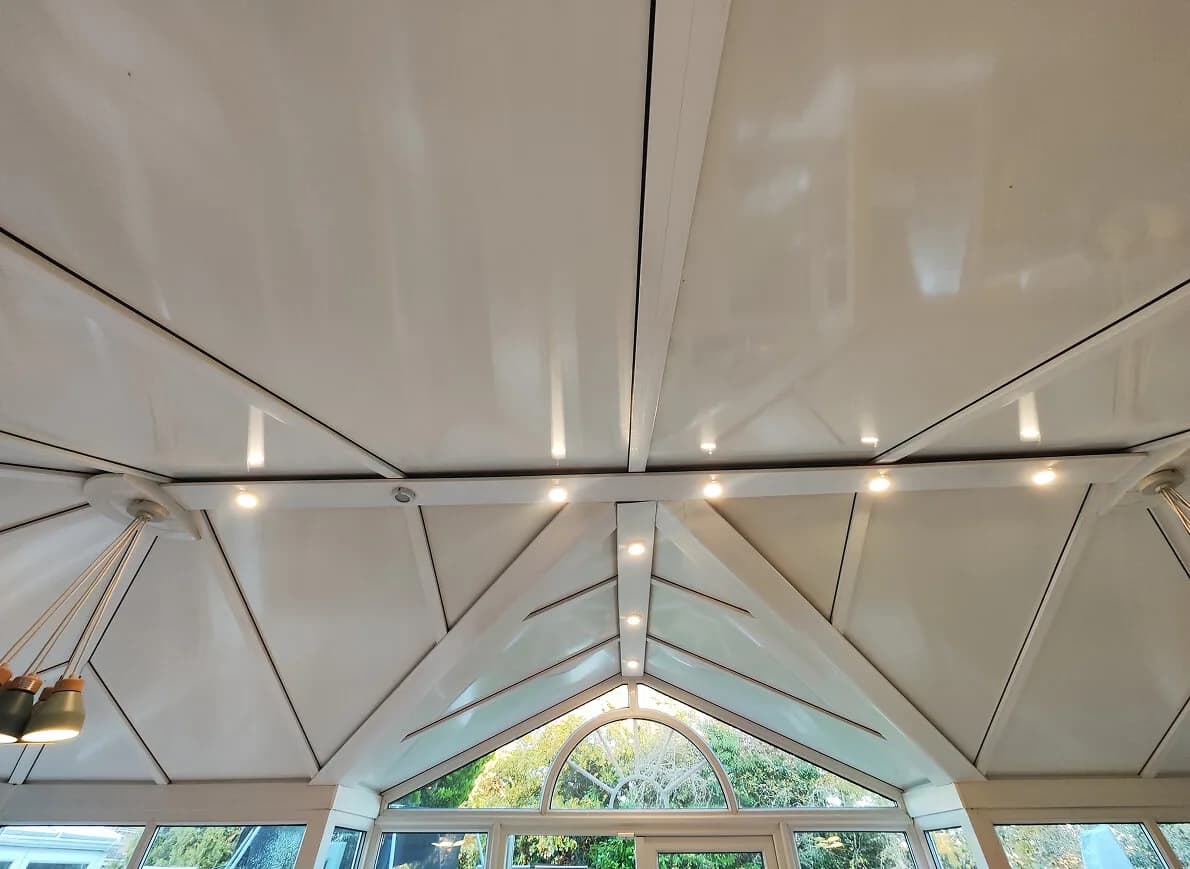 The underside of an insulated panel conservatory roof from inside the room, a hipped white ceiling with recessed spotlights along the glazing bars.